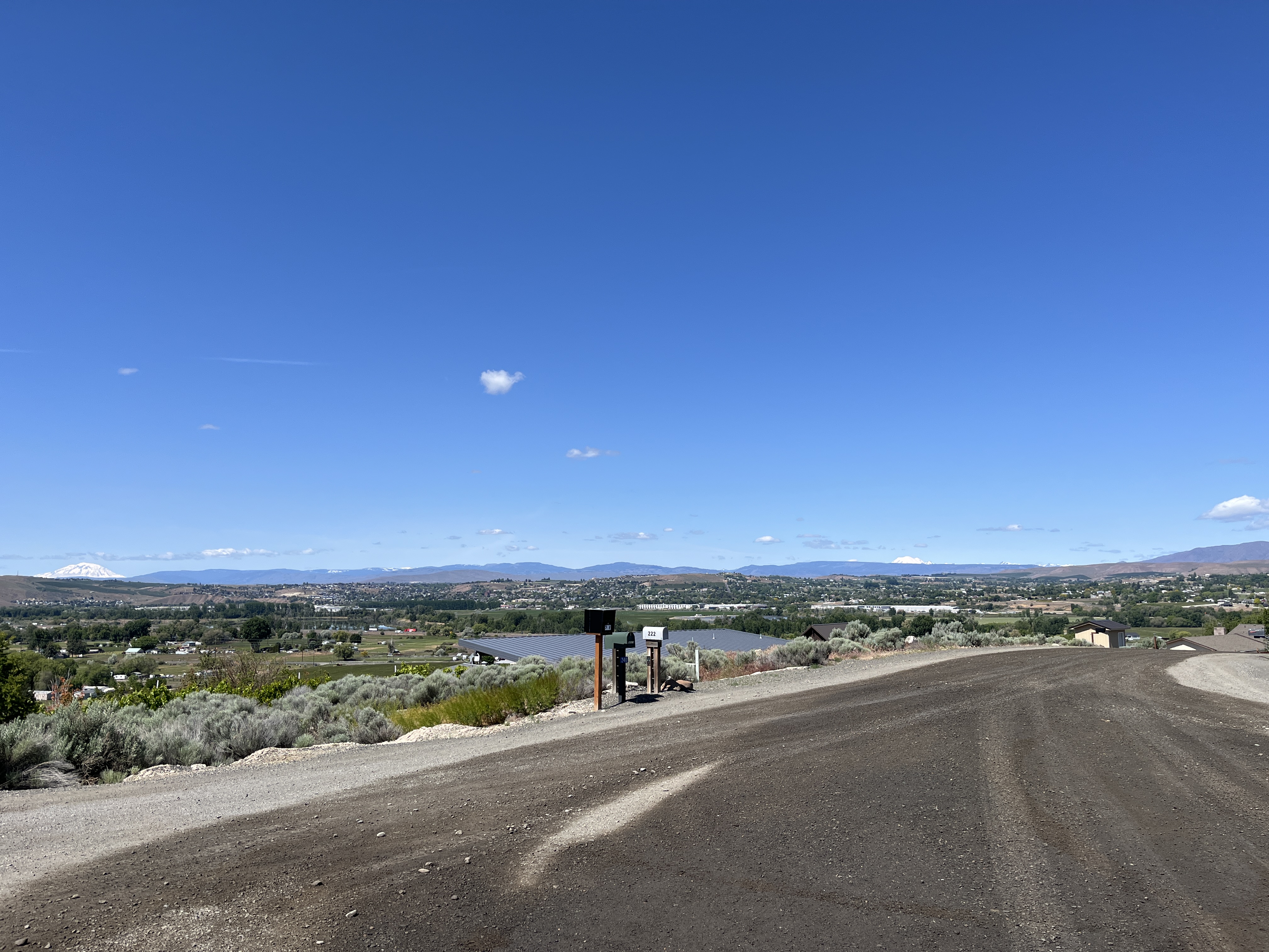 Una carretera de gravilla con tres buzones y vista de un valle con campos y edificios. Al fondo puede verse una cadena de montañas bajo un cielo azul y soleado.